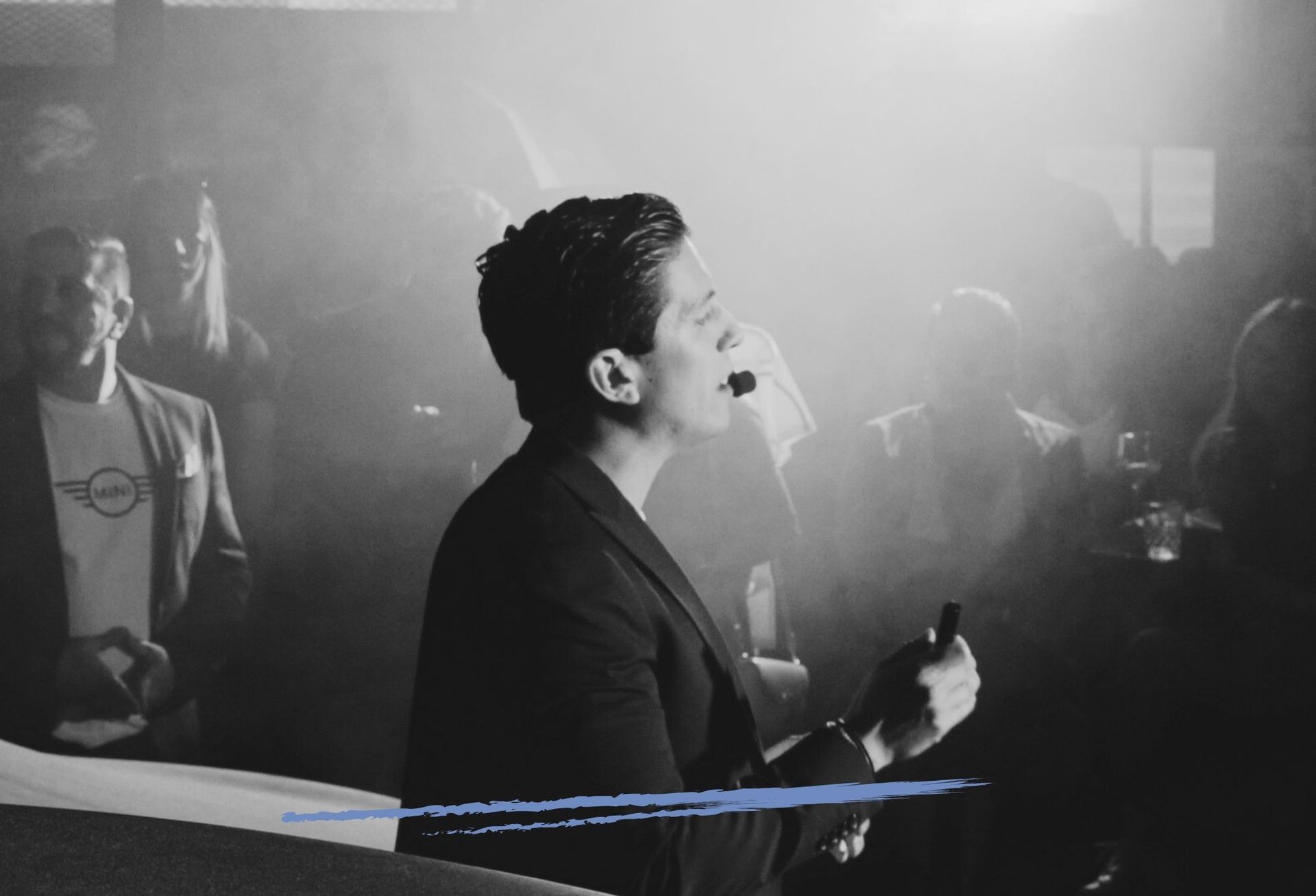 Person speaking into a headset microphone in a smoky room, audience blurred in the background.